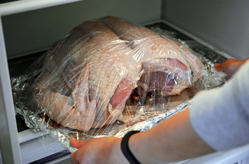 A salted, dry-brined Spatchcock Turkey is loosely covered with plastic wrap and placed in the fridge.