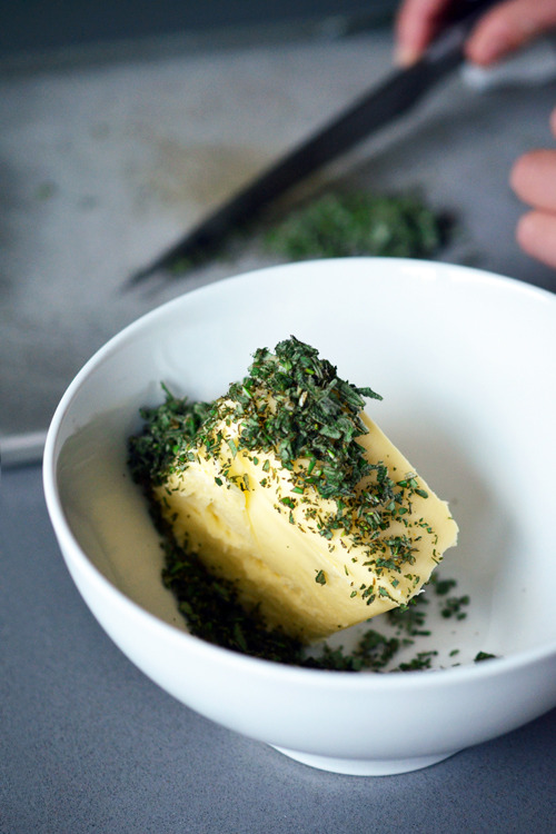 Adding the minced fresh herbs to a block of butter in a white bowl.