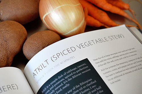 The Paleo By Season cookbook is opened up to the Atkilt (Ethiopian Spiced Vegetable Stew) recipe on a table with potatoes, onion, and carrots.