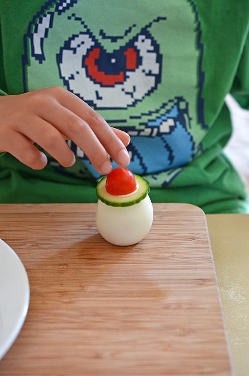 Someone putting a sliced cucumber and grape tomato on top of a hard boiled egg for crazy clown eggs.