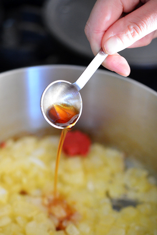 Someone pouring a teaspoon of fish sauce into a pot of onions and tomato paste for umami gravy.
