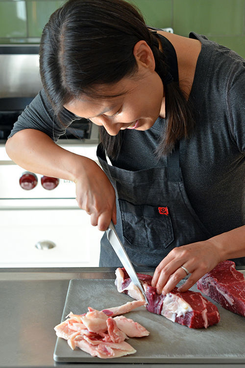 A woman is trimming the fat off of a boneless New York strip steak with a chef's knife. 