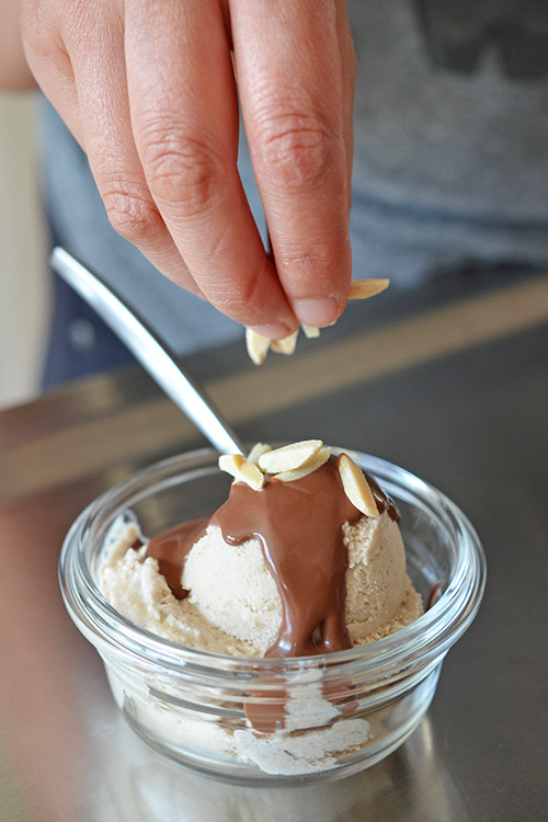 A hand is adding toasted almonds to the top of a scoop of Dairy-Free Vanilla Ice Cream with chocolate Ganache 