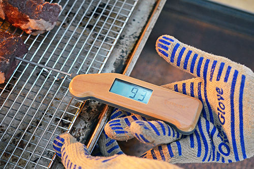 A gloved hand is checking the temperature of the strip steaks with a Thermapen Mk4. 