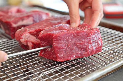 A metal probe of a meat thermometer is being inserted into a raw strip steak on a wire rack in a rimmed baking sheet.