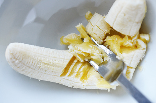 A medium banana is mashed with a fork in a white bowl.