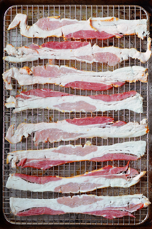 A rimmed baking sheet with eight strips of raw bacon on a stainless steel wire rack.
