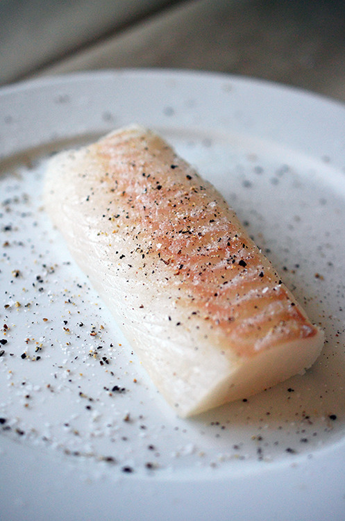 A cod fillet seasoned with salt and pepper on a white plate.