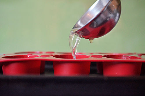 A ladle is pouring cooled bone broth into a red silicone ice mold in a rimmed baking sheet.