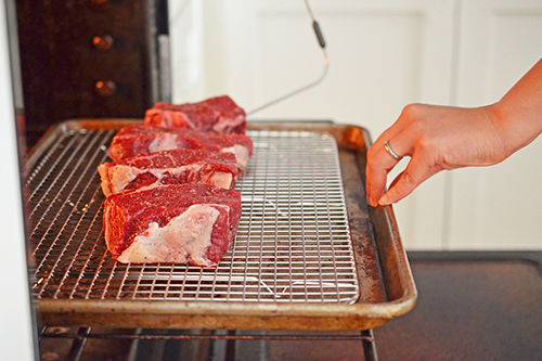 Placing the four strip steaks into the oven on a wire rack in a rimmed baking sheet. One of the steaks has a thermometer probe inserted inside it.