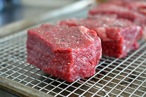 A side shot of seasoned strip steaks on a wire rack in a rimmed baking sheet.