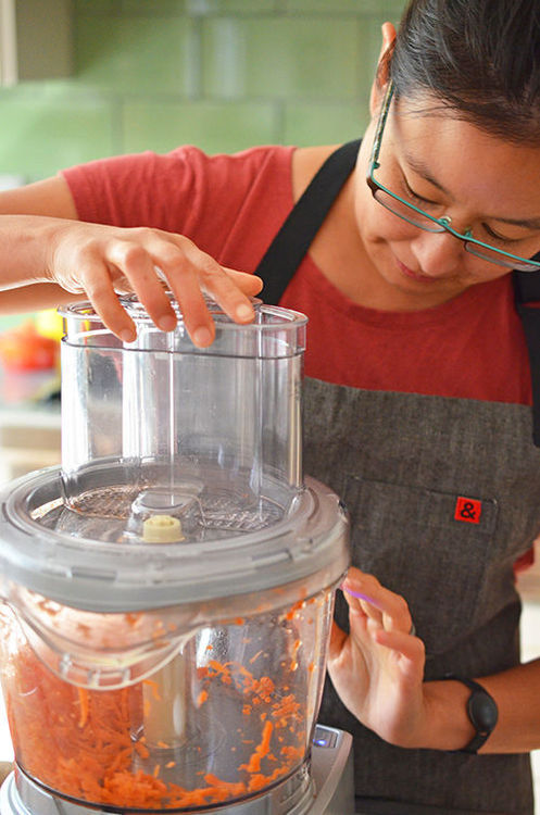 A woman is grating carrots in a food processor to make Paleo Pumpkin and Carrot Muffins 