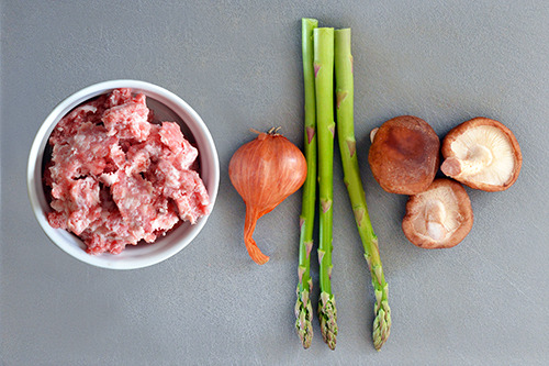 An overhead shot of raw ground pork, a shallot, asparagus, and three shiitake mushrooms.