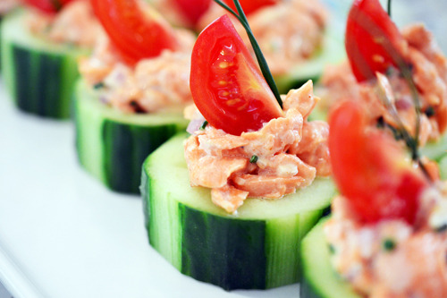 Two rows of the cucumber spicy salmon bites garnished with a slice of tomato on top with a piece of chive.