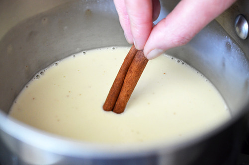 A pot of coconut milk coming to a simmer, and someone adding a cinnamon stick.
