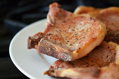 Seared pork chops sitting on a white plate for Bacon Apple Smothered Pork Chops.