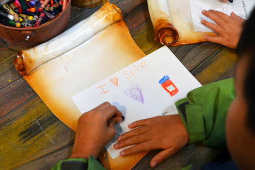 A child drawing on a piece of paper with a crayon writing, "I love spice."