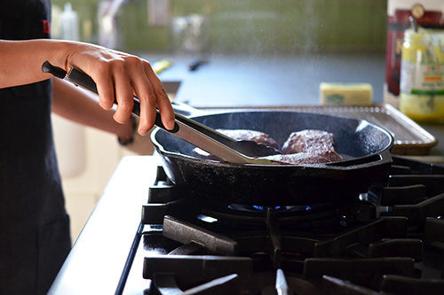 Side shot of someone frying steaks in a large cast iron skillet.