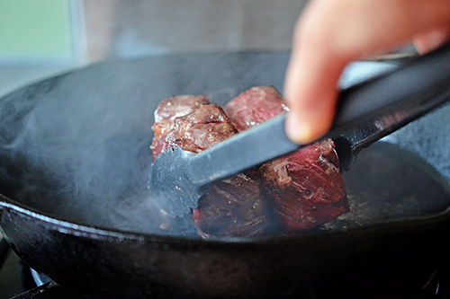 A pair of tons is holding two strip steaks on their sides to brown them in a cast iron skillet.