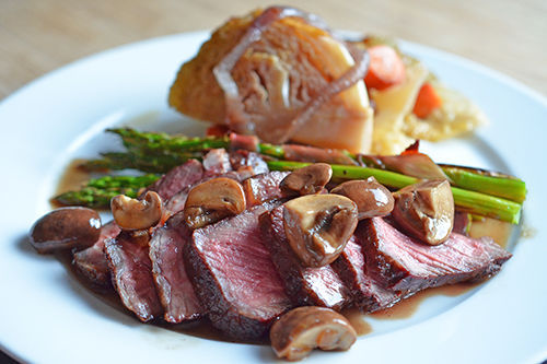 A side shot of a white plate with sliced medium rare steak topped with mushrooms. There are asparagus spears and braised cabbage on the plate as well.
