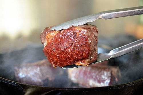 A pair of tons is holding a golden brown strip steak on top of a cast iron skillet. 