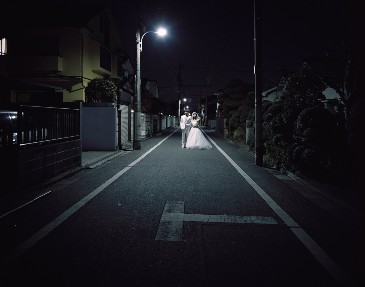 A couple wearing wedding dress and tuxedo is standing on the street at night.