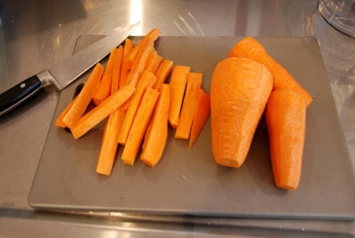 Carrots peeled and cut length-wise sitting on a cutting board with a knife resting on the cutting board.