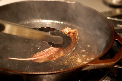 Someone searing a lamb chop in a cast iron skillet.