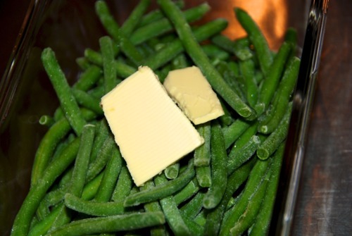 A shallow microwaveable dish fill with green beans with a thin slab of butter on top.