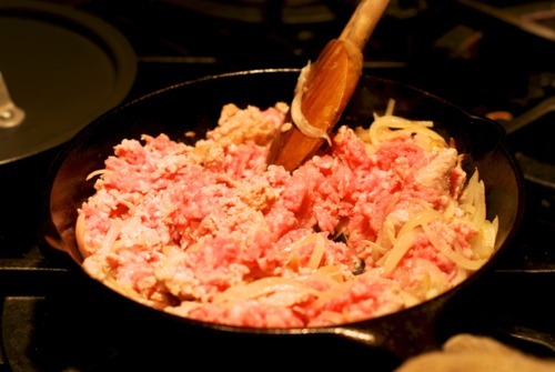Someone cooking ground pork in a cast iron skillet with a wooden spoon.