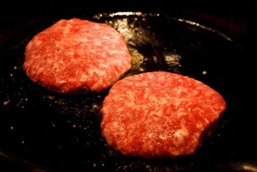 Two New Zealand lamb burgers frying on a cast iron skillet.