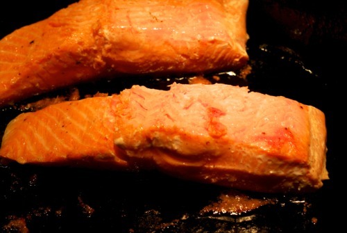 Two salmon fillets searing in a cast iron skillet.