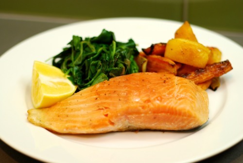 A cooked sous vide wild king salmon fillet on a plate with a lemon wedge, sautéed spinach, and carrots.