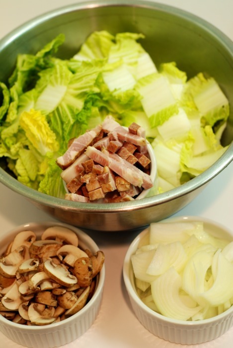 Assembled ingredients for stir fried napa cabbage. Chopped lettuce, chopped bacon, sliced mushrooms, and sliced onions all sit in separate bowls on a counter.