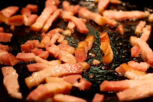 Tiny pieces of bacon are being fried in a cast iron skillet.