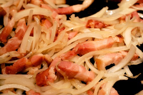 Tiny pieces of bacon are being fried in a cast iron skillet with thinly sliced onions.