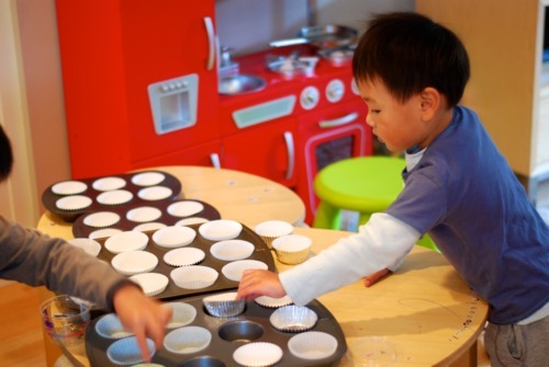 Two little boys placing muffin liners in cupcake trays.