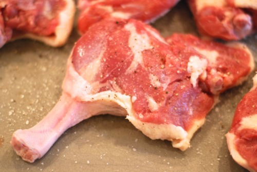 Duck legs seasoned with salt and pepper on a cutting board.