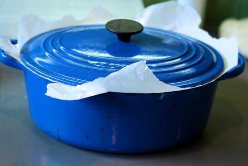 A blue dutch oven sits on a counter top with parchment paper on top of the bottom of the dutch oven and the lid on top of it.