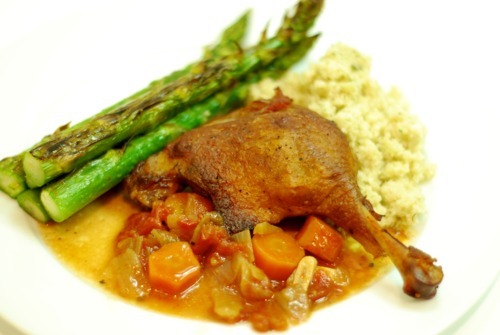A plate containing crispy braised duck legs on top of a vegetable mixture of braised carrots and onions. On the side is also asparagus and quinoa.