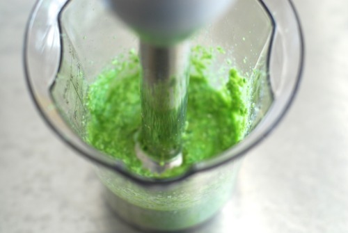 Coconut milk, celery, and cilantro being mixed in a cup by an immersion blender.