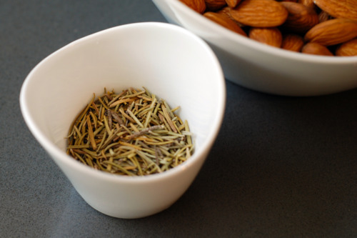 A small bowl of dried rosemary next to a large bowl of almonds.