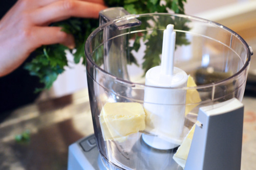 Slices of butter in a food processors, with someone adding in parsley.