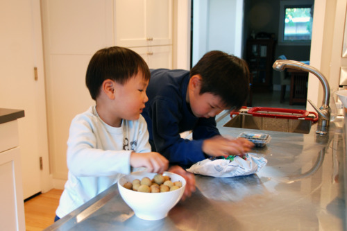 Two young children peeling the skins of the longan fruit in a kitchen.