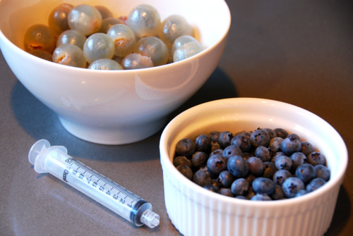 A bowl of peeled longan and a bowl of blueberries on a countertop with a syringe to make bloody eyeballs.