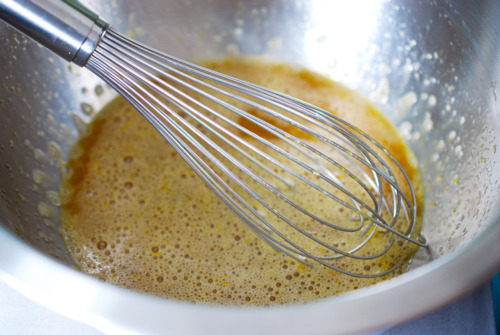 A whisk is mixing eggs and maple syrup in a large metal bowl.