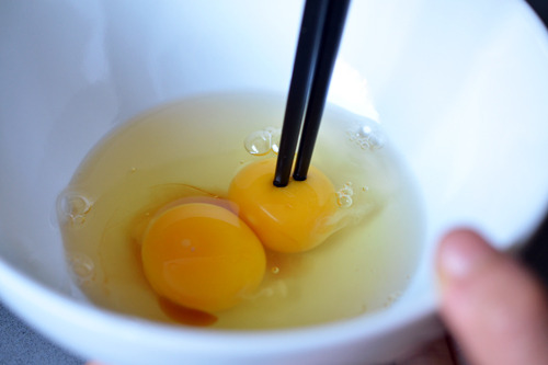 Someone puncturing the yolks of the cracked eggs in a bowl with chopsticks.