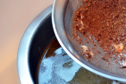 A mesh sieve has bone pieces inside and it is hovering over a bowl of beef bone broth.